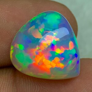 Könnte beinhalten: Ein polierter, dreieckiger Opal-Edelstein mit einer lebendigen Farbgebung. Der Opal hat eine durchscheinende Basis mit internen Blitzen in Orange, Gelb, Grün und Blau, wodurch ein Farbspiel-Effekt entsteht. Der Stein ist glatt und abgerundet.