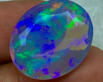 May include: An oval opal gemstone with a faceted surface. The stone exhibits a vibrant play of colour, including shades of blue, green, purple, and orange. The gemstone is held between fingers, showcasing its polished appearance and iridescent qualities.