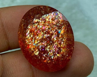 May include: A large, oval-shaped, iridescent sunstone cabochon with a deep orange base and a scattering of gold, red, and green glitter.