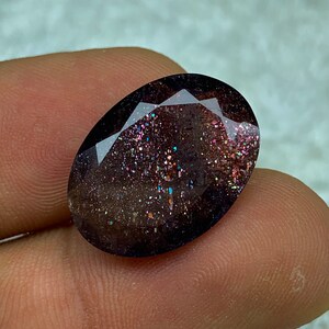 May include: A dark gray oval-shaped gemstone with a sparkly, iridescent finish. The gemstone has a speckled appearance with hints of pink, blue, and green.