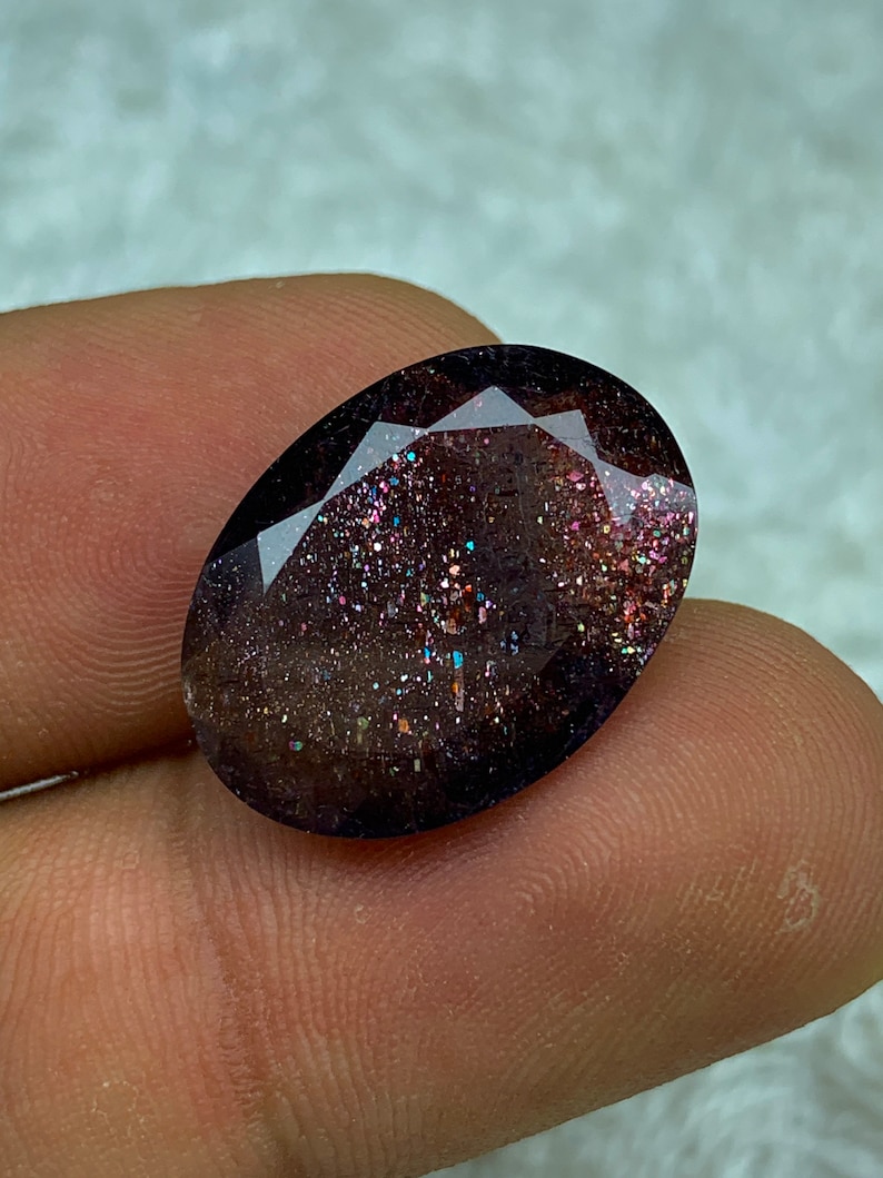 May include: A dark gray oval-shaped gemstone with a sparkly, iridescent finish. The gemstone has a speckled appearance with hints of pink, blue, and green.