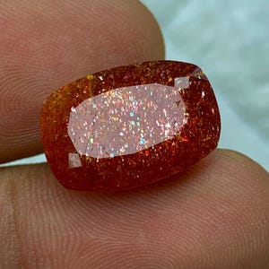 May include: A large, rectangular, orange sunstone gemstone with a shimmering, iridescent finish. The stone has a reddish-brown hue with a subtle sparkle.