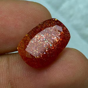 May include: A red, orange and gold sunstone gemstone with a rectangular shape and a sparkly, iridescent finish.