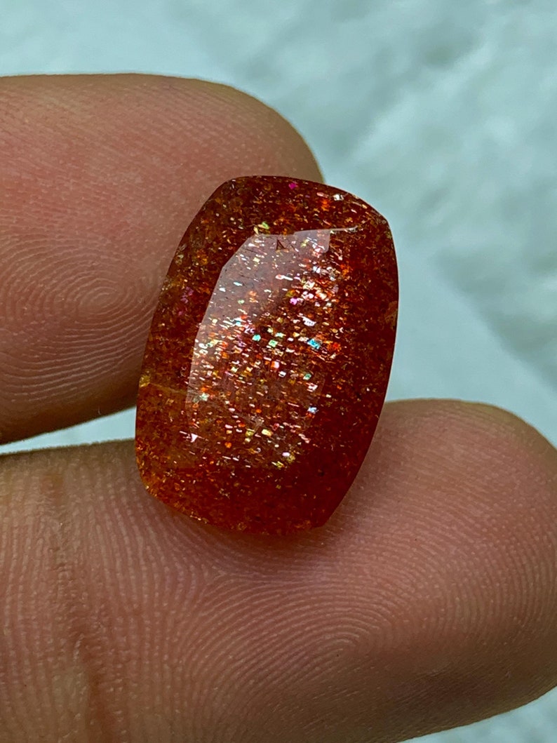May include: A red, orange and gold sunstone cabochon with a smooth, polished surface. The stone has a shimmering, iridescent quality.