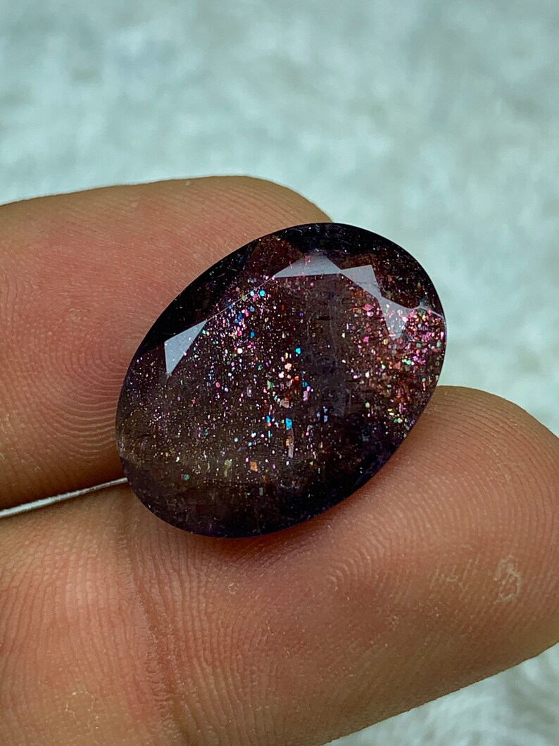 May include: A dark purple oval gemstone with iridescent glitter embedded throughout. The gemstone has a faceted cut.