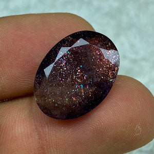 May include: A dark purple oval-shaped gemstone with a sparkly, iridescent finish. The stone has a unique, speckled appearance with flecks of gold, silver, and pink.