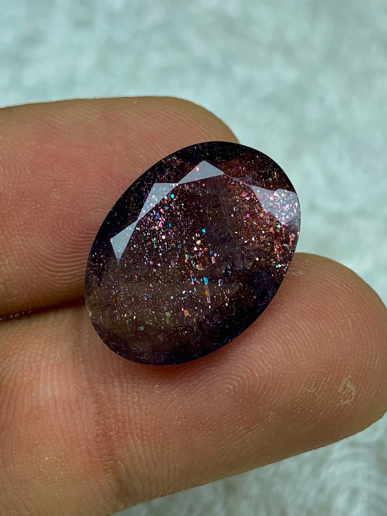 May include: A dark purple oval-shaped gemstone with a sparkly, iridescent finish. The stone has a unique, speckled appearance with flecks of gold, silver, and pink.