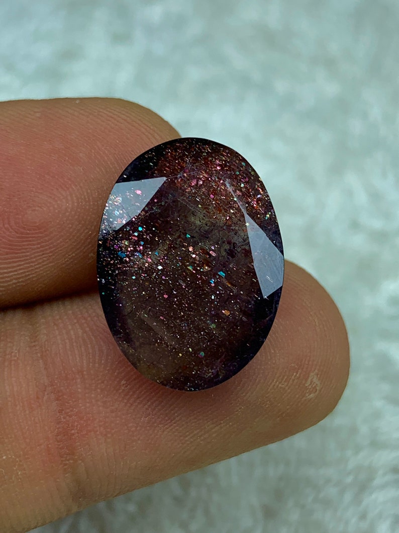 May include: A dark purple oval-shaped gemstone with a sparkly, iridescent finish. The stone has a speckled appearance with flecks of gold, silver, and pink.