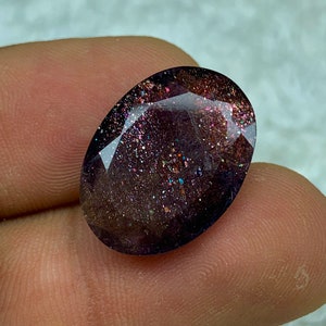 May include: A dark purple oval gemstone with iridescent glitter embedded throughout. The gemstone is cut and polished.
