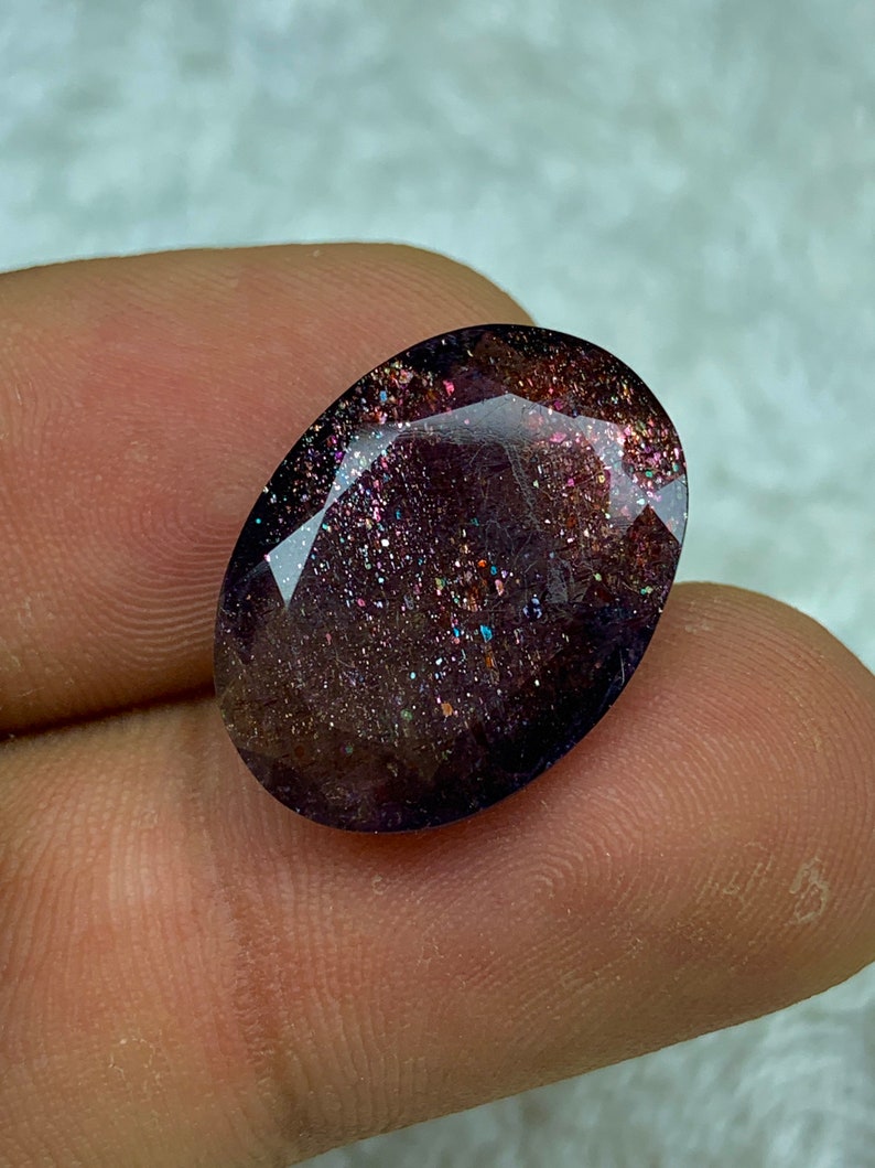 May include: A dark purple oval gemstone with iridescent glitter embedded throughout. The gemstone is cut and polished.