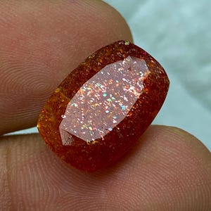 May include: A large, reddish-orange sunstone gemstone with a faceted cut. The stone has a shimmering, iridescent quality with a rainbow of colours.