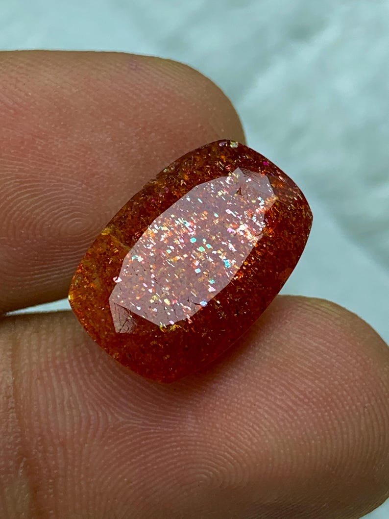 May include: A large, reddish-orange sunstone gemstone with a faceted cut. The stone has a shimmering, iridescent quality with a rainbow of colours.