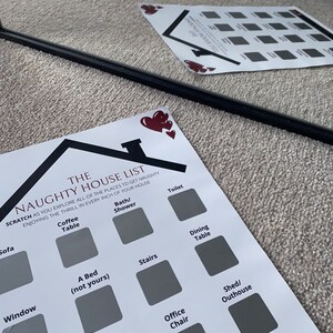May include: A white paper with the text "The Naughty House List" and scratch-off squares. The list includes locations like "Sofa," "Coffee Table," and "A Bed (not yours)." A black, thin, straight object is in the foreground.
