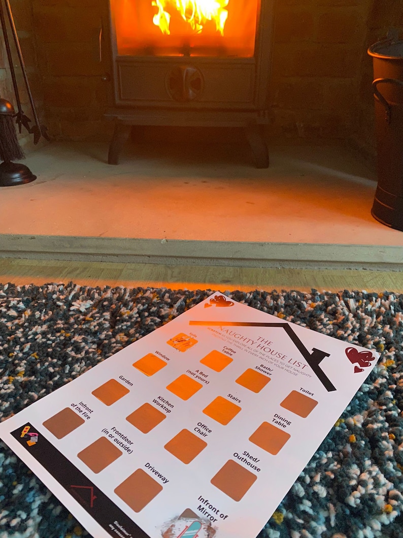 May include: A printed "Naughty House List" sits on a colorful rug in front of a fireplace. The list features various locations like the kitchen, stairs, and front door, with scratch-off areas. A cozy scene with a burning fire.