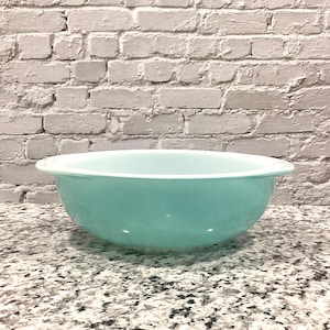 May include: A turquoise mixing bowl with a white rim. The bowl is made of a smooth material and has a rounded shape. The background is a light gray brick wall. The bowl is sitting on a speckled countertop.