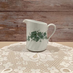 May include: A white ceramic creamer with a handle and spout. The creamer is decorated with a green leaf pattern and sits on a white lace doily. The background is a wooden wall.