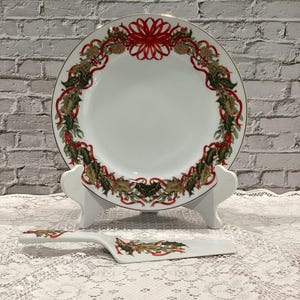 May include: White ceramic plate and cake server set with a Christmas wreath design. The plate has a gold rim and a wreath of holly, pinecones, and red ribbons. The cake server mirrors the plate's design.