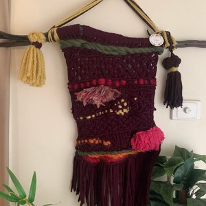 Puede incluir: Un tapiz de macramé de color granate con detalles de hilo de colores, incluyendo rosa, amarillo y verde. El tapiz tiene un borde de flecos y está sujeto a una rama con cuerda.