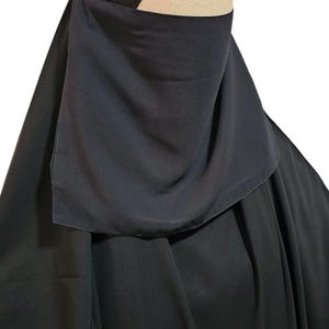 May include: A black niqab, a traditional Islamic veil, featuring a head covering and face veil. The fabric drapes over the shoulders. The niqab is designed to cover the face, leaving only the eyes visible. The head covering is a solid, structured design.