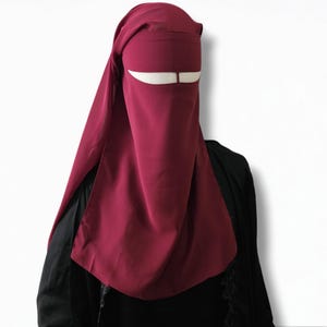 May include: A burgundy niqab with a white eye opening. The niqab is made of a soft, flowing fabric.