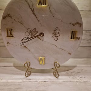 Puede incluir: Un reloj de pared redondo con un diseño de mármol blanco y dorado. La esfera del reloj presenta números romanos dorados y motivos decorativos de flores y mariposas. El reloj está sostenido por un soporte de metal dorado.