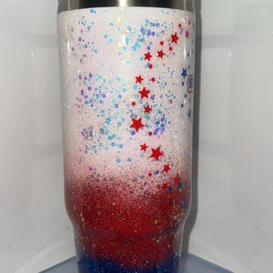 May include: A tall cylindrical tumbler with a white lid and a clear straw. The body features a glittery gradient transitioning from white at the top to red in the middle and blue at the bottom, decorated with small red star-shaped accents.
