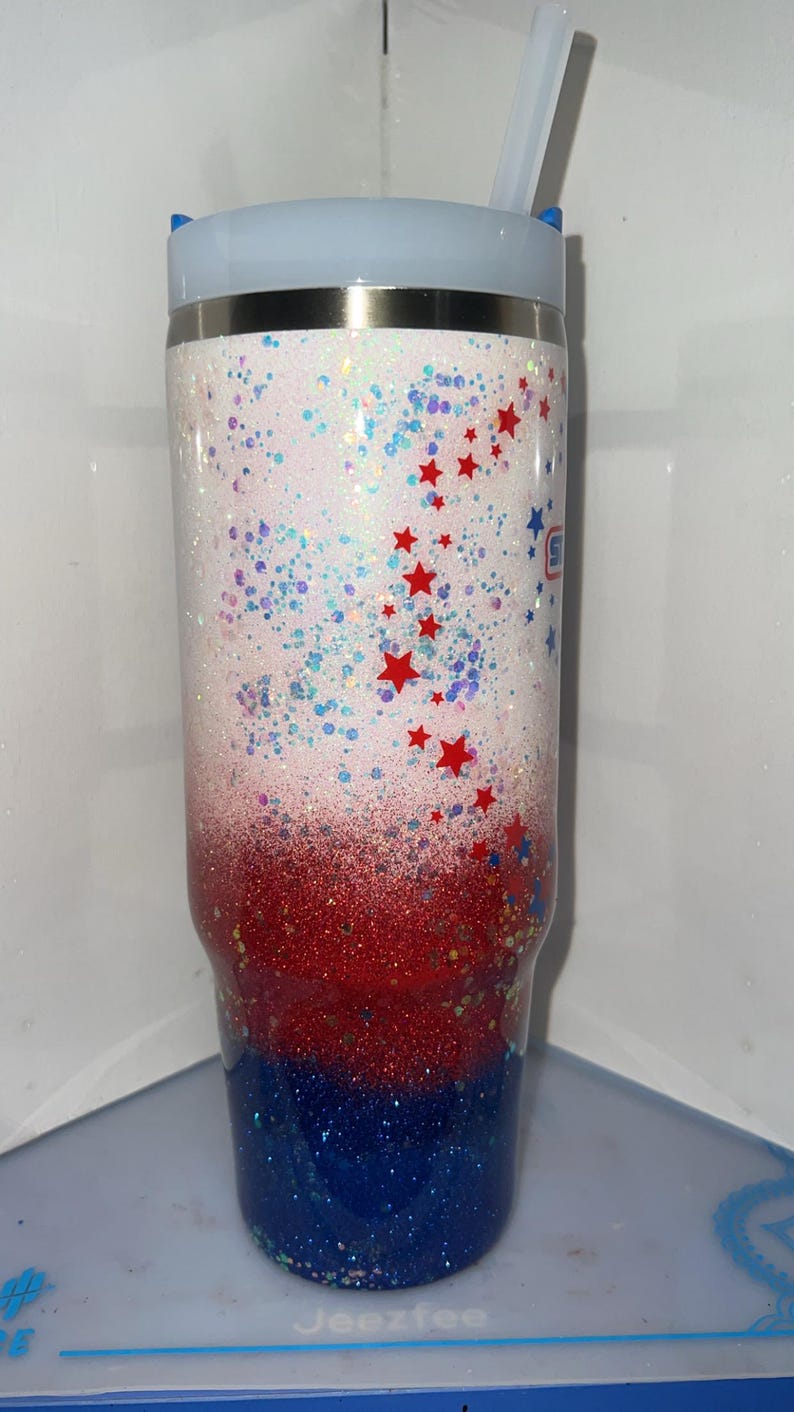 May include: A tall cylindrical tumbler with a white lid and a clear straw. The body features a glittery gradient transitioning from white at the top to red in the middle and blue at the bottom, decorated with small red star-shaped accents.