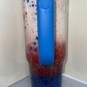 May include: A tall insulated tumbler with a handle, featuring a glittery gradient design that transitions from white at the top to red in the middle and dark blue at the bottom. The surface is decorated with small red and blue star-shaped accents.