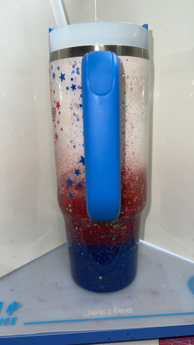 May include: A tall insulated tumbler with a handle, featuring a glittery gradient design that transitions from white at the top to red in the middle and dark blue at the bottom. The surface is decorated with small red and blue star-shaped accents.