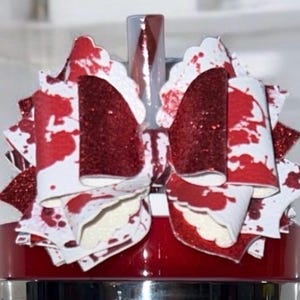 May include: A decorative hair bow with a white base and red splatter design. The bow features a glittery red center and accents, creating a striking contrast. The bow is attached to a red base.