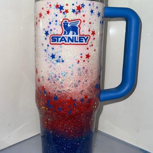 May include: A tall insulated tumbler with a handle, featuring a white, red, and blue glitter gradient finish. The cup is decorated with small red and blue stars and displays the Stanley logo in blue and red text on the upper white section.