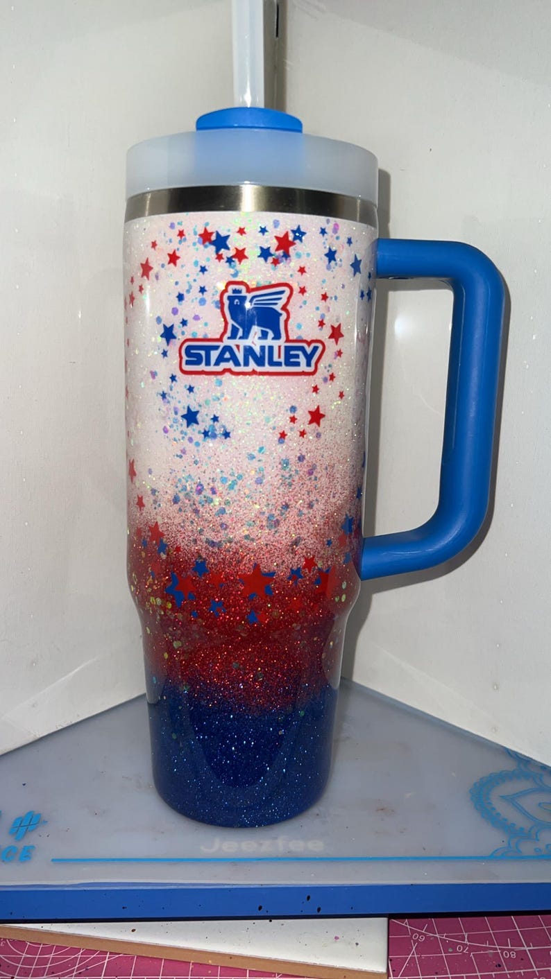 May include: A tall insulated tumbler with a handle, featuring a white, red, and blue glitter gradient finish. The cup is decorated with small red and blue stars and displays the Stanley logo in blue and red text on the upper white section.
