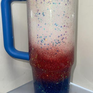 May include: A tall stainless steel tumbler featuring a glittery ombre finish that transitions from white at the top to red in the middle and dark blue at the base. The cup includes a bright blue handle and a matching blue lid with a clear straw opening.