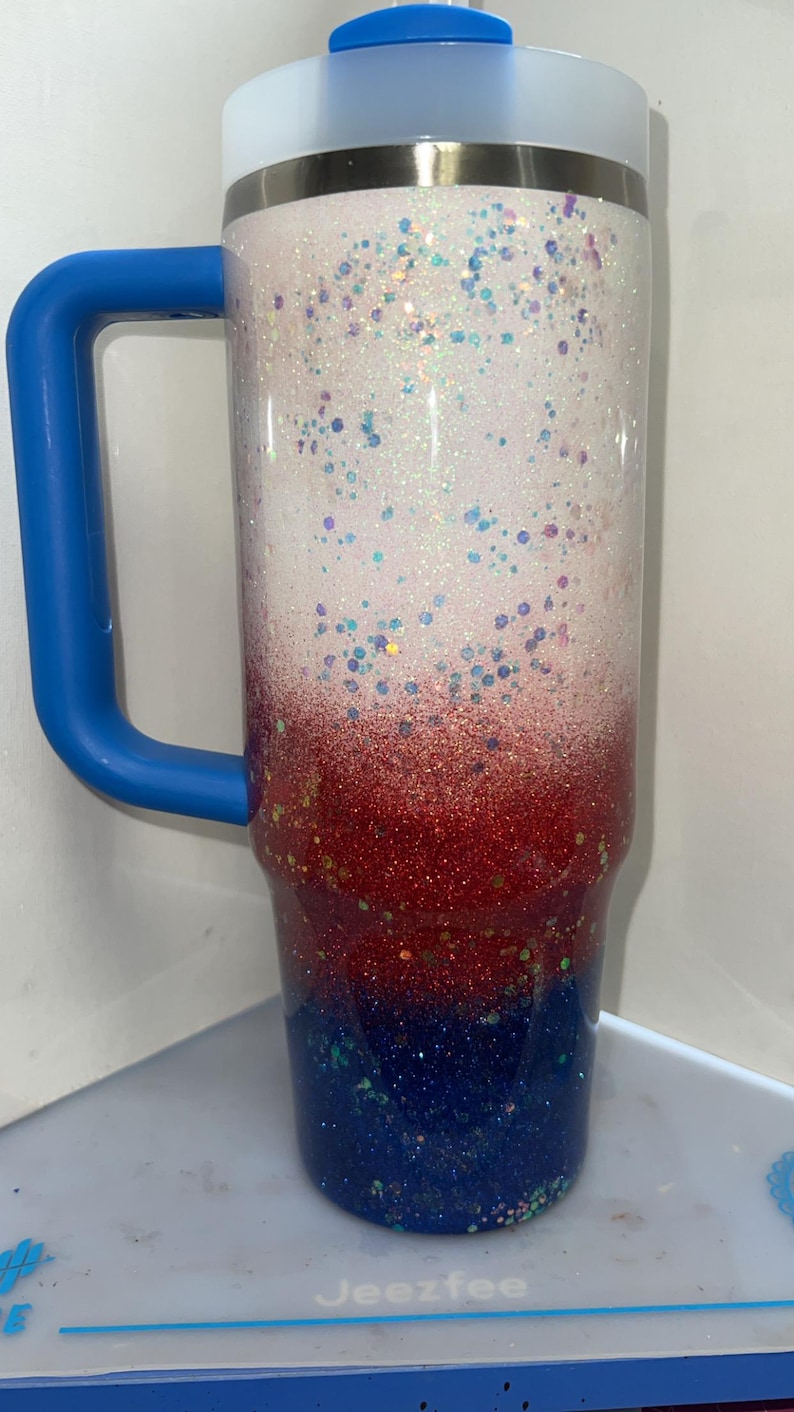 May include: A tall stainless steel tumbler featuring a glittery ombre finish that transitions from white at the top to red in the middle and dark blue at the base. The cup includes a bright blue handle and a matching blue lid with a clear straw opening.
