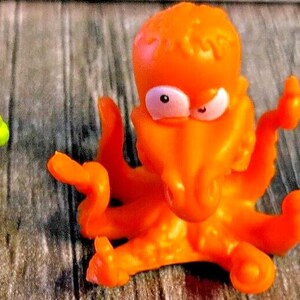 May include: Three colorful, cartoonish toys are displayed. A green creature with multiple eyes and a white bandage, an orange octopus, and a white creature with red accents and green legs. The toys are on a wooden surface.