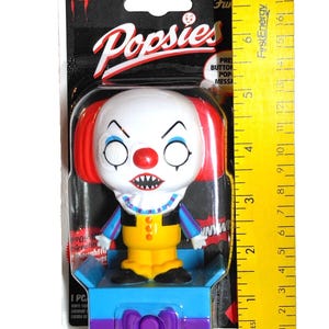 May include: A Funko Popsies vinyl figure of Pennywise the clown from the movie "It". The figure has a white face with red hair, a yellow outfit, and a blue base. The packaging includes the text "Popsies".