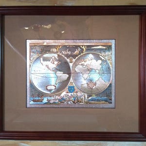 May include: Framed vintage world map print. The map features two hemispheres with detailed illustrations of continents, oceans, and historical elements. The print is set within a brown mat and a dark brown wooden frame, suitable for home decor.
