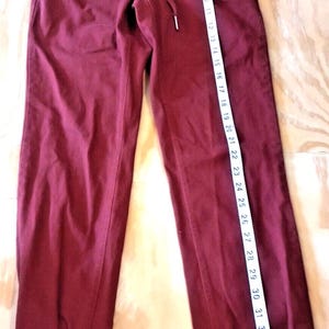 May include: Burgundy-colored sweatpants with an elastic waistband and drawstring. The pants have ribbed cuffs at the ankles. A measuring tape shows the length of the pants to be approximately 36 inches.