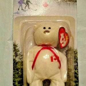 May include: A white plush bear toy named "Maple" with a red ribbon and a Canadian flag. The bear is in a clear plastic package with a "Ty" heart-shaped tag. The packaging has the text "Maple The Bear".