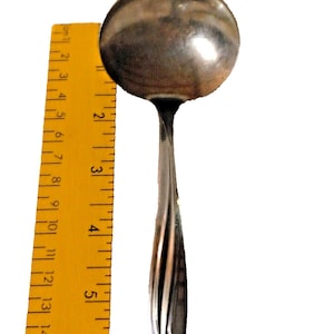 May include: A stainless steel ladle with a rounded bowl and a long, curved handle. The ladle is positioned next to a yellow ruler with black markings. The ruler shows measurements in inches.