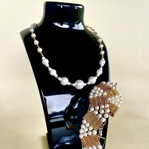 May include: A white pearl necklace and a gold and white pearl bracelet on a black jewelry display stand.