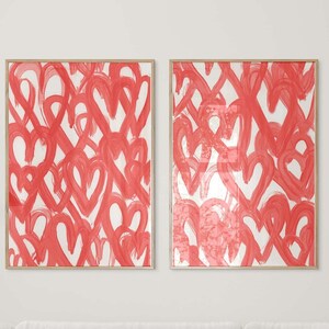 May include: Two framed abstract art prints with a white background and a pattern of red hearts. The prints are hanging on a white wall above a white sofa.