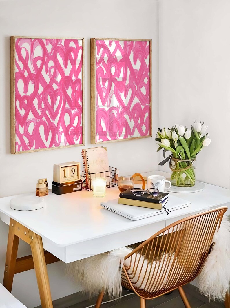 May include: A white desk with a wooden frame and a gold metal chair. There are two framed pink heart prints on the wall above the desk. A laptop, books, a candle, a calendar, and a vase of white tulips are on the desk.