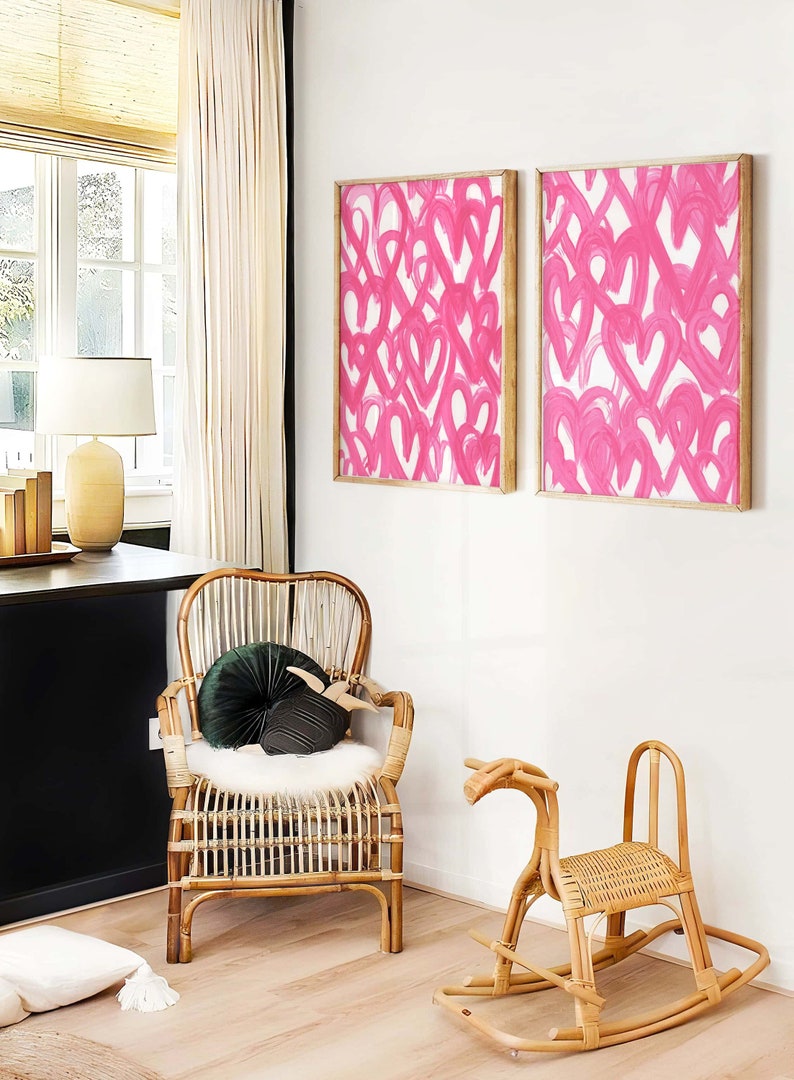 May include: Two framed pink abstract paintings with a white background and a wooden frame. A wicker rocking horse and a wicker chair with a green velvet pillow are in the foreground.