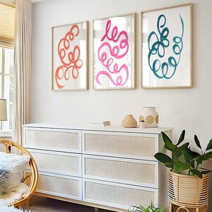 K&ouml;nnte beinhalten: Drei gerahmte abstrakte Kunstdrucke mit bunten, wirbelnden Linien. Die Drucke h&auml;ngen an einer wei&szlig;en Wand &uuml;ber einer wei&szlig;en Kommode mit geflochtenen Korb-Schubladen.