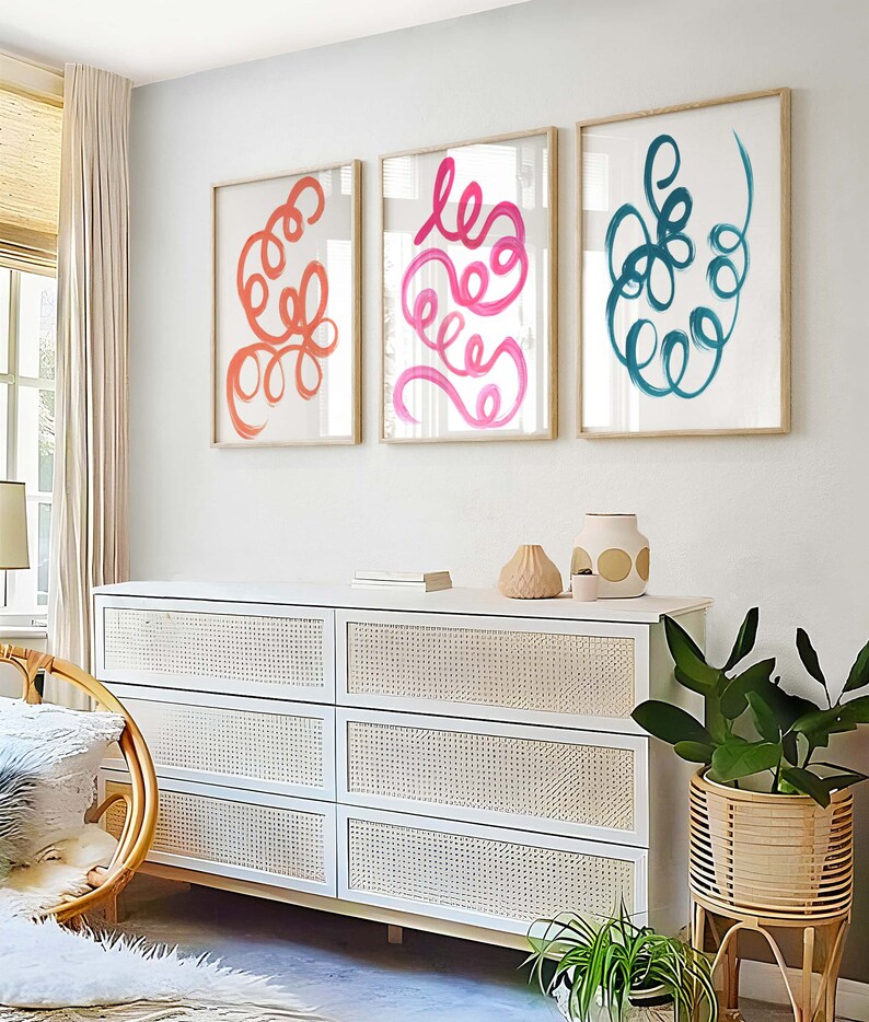 K&ouml;nnte beinhalten: Drei gerahmte abstrakte Kunstdrucke mit bunten, wirbelnden Linien. Die Drucke h&auml;ngen an einer wei&szlig;en Wand &uuml;ber einer wei&szlig;en Kommode mit geflochtenen Korb-Schubladen.