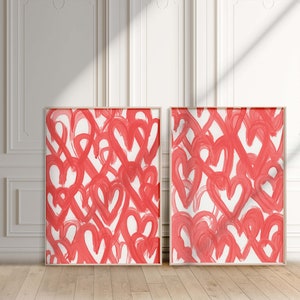 May include: Two framed abstract paintings with a white background and a pattern of red hearts. The paintings are hanging on a white wall.