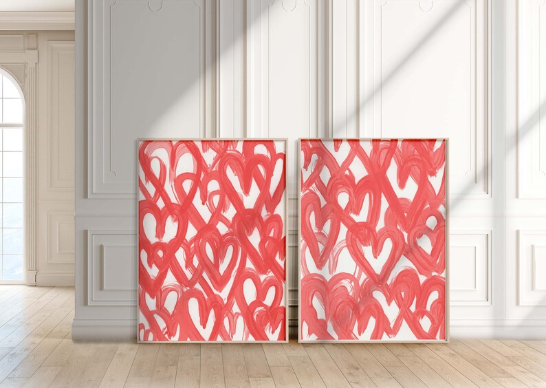 May include: Two framed abstract paintings with a white background and a pattern of red hearts. The paintings are hanging on a white wall.