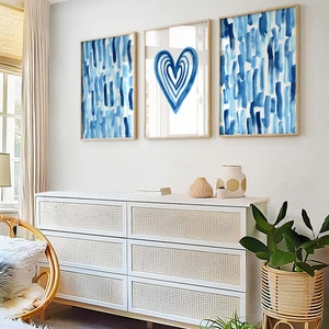 Puede incluir: Tres impresiones art&iacute;sticas abstractas enmarcadas. Dos impresiones presentan pinceladas azules verticales sobre un fondo blanco. La impresi&oacute;n central muestra una forma de coraz&oacute;n azul sobre un fondo blanco. Las impresiones se exhiben sobre una c&oacute;moda blanca con frentes de cajones tejidos.