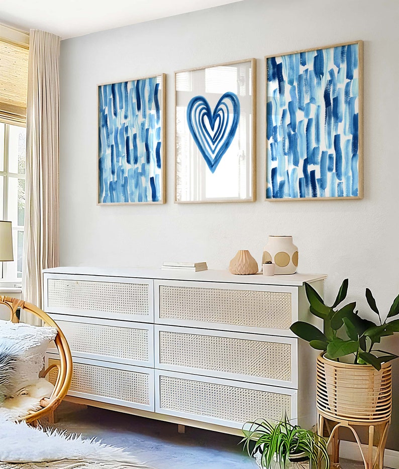 Puede incluir: Tres impresiones art&iacute;sticas abstractas enmarcadas. Dos impresiones presentan pinceladas azules verticales sobre un fondo blanco. La impresi&oacute;n central muestra una forma de coraz&oacute;n azul sobre un fondo blanco. Las impresiones se exhiben sobre una c&oacute;moda blanca con frentes de cajones tejidos.